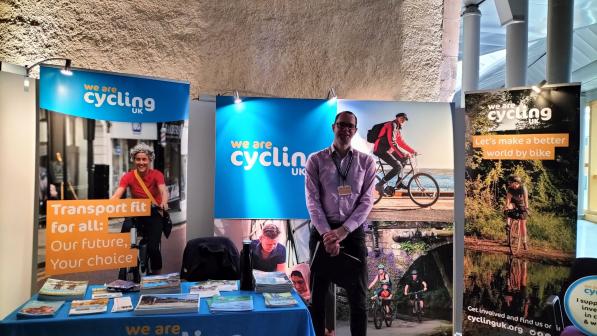 A man in shirt and trousers with a name tag round his neck and holding a clipboard. He is at an exhibition stand with Cycling UK branded posters and table