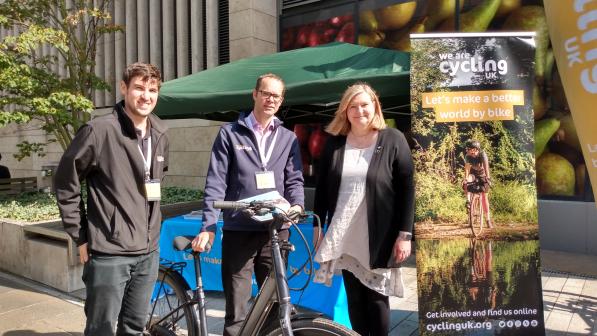Three people (two men and a woman) are standing in front of a gazebo with a Cycling UK banner to the side. The man in the centre is holding an e-bike.