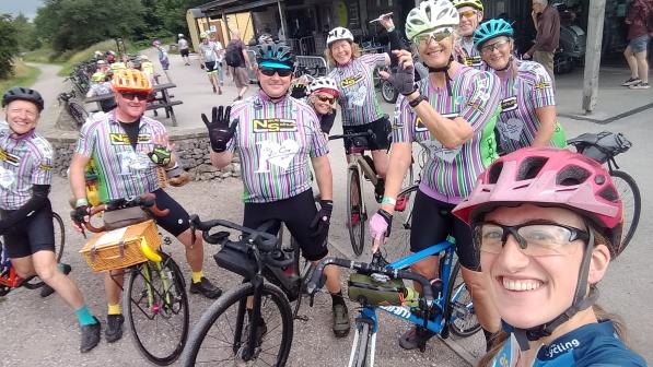 A group of people all wearing cycling kit and cycle helmets are standing with their bikes and smiling at the camera