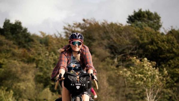 Jo Lankester is cycling along a wide flat grassy track wearing cycling glasses and a red checked shirt. There is a woodland behind her