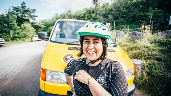 Jo Shwe is smiling wearing a green cycle helmet and standing in front of a yellow van which is parked on the side of a tree lined road
