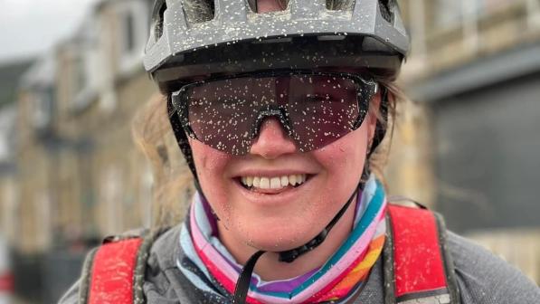 Jodie Brumhead is pictured wearing a black helmet and sunglasses. She is smiling and her cheeks are flushed and there are small flecks of mud covering her face and top