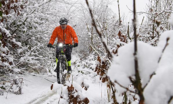 A man is cycling along a snow covered cycle track. Snow covered trees are on both sides of the track. The cyclist is wearing an orange coat and cycle helmet and riding a mountain bike