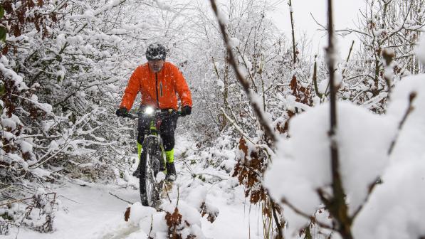 A man is cycling along a snow covered cycle track. Snow covered trees are on both sides of the track. The cyclist is wearing an orange coat and cycle helmet and riding a mountain bike