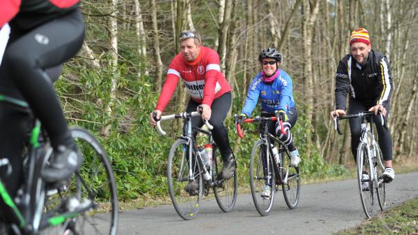 A group of people are cycling along a tarmac lane through a wood. They are all on road bikes and wearing cycling kit. They are smiling.