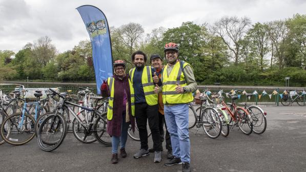 Three adults wearing hi-vis vests and bike helmets stand smiling in front of a row of bicycles at an outdoor cycling event. A blue flag with a cycling logo is visible behind them, with trees in the background under an overcast sky.