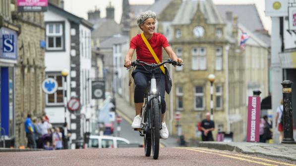 A woman with grey curly hair wearing a bright red top cycles up a hill towards the camera on a high street