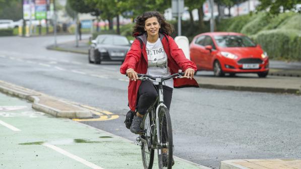 A woman with dark brown hair and a red coat rides along a separated cycle lane beside a road