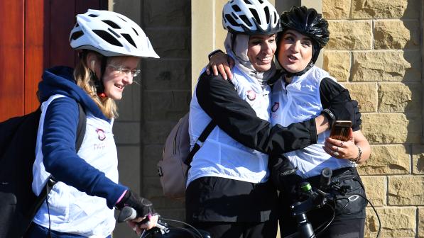 Three women in cycling helmets and white vests share a joyful moment outside a stone building. Two of them hug and smile while holding a phone, the third stands nearby with a bike, also smiling.