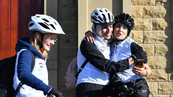 Three women in cycling helmets and white vests share a joyful moment outside a stone building. Two of them hug and smile while holding a phone, the third stands nearby with a bike, also smiling.