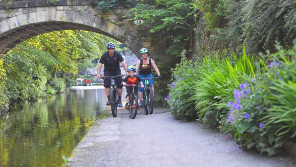 Two adults and a child cycling along a canal. They are all wearing tshirts and it is sunny.