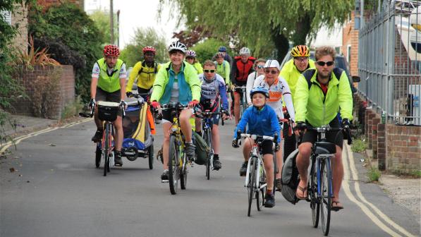 The image shows a diverse group of cyclists, including adults and children, riding along a quiet street lined with greenery. Many participants are wearing bright safety gear, such as fluorescent jackets and helmets. Some are riding traditional bikes, while others have cargo bikes or bicycles with child trailers, reflecting a family-friendly and inclusive cycling event. The atmosphere appears lively and community-oriented, with participants smiling and enjoying the ride.