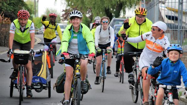 A mixed group of people is cycling along an urban road. There are different kinds of bikes and even a trailer with kids in. They are smiling and waving. Some are wearing hi-vis but most are in normal clothing. 