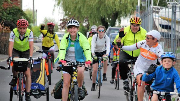 A mixed group of people is cycling along an urban road. There are different kinds of bikes and even a trailer with kids in. They are smiling and waving. Some are wearing hi-vis but most are in normal clothing. 