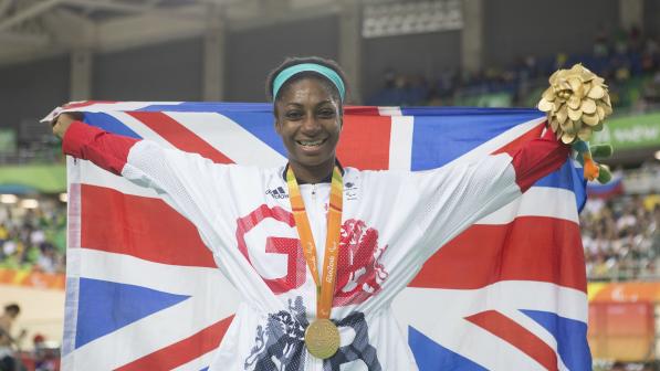 Kadeena Cox is holding the Union Jack flag behind her and is wearing a gold medal that has the words Rio 2016 along the ribbon. She is also holding gold flowers and is pictured inside a stadium