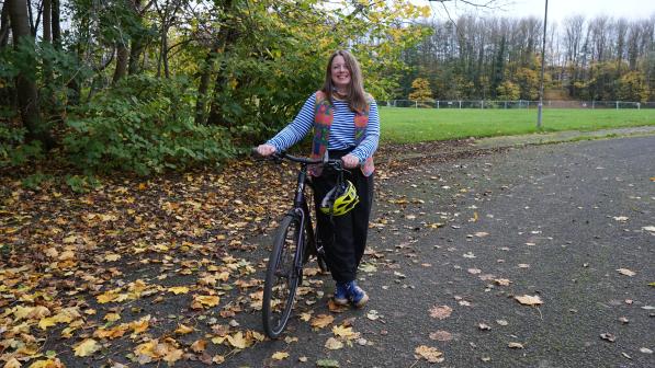Katherine Cory, of Women on Wheels, stands smiling beside her bike on a leaf-covered path in a park, with trees and a football pitch in the background.