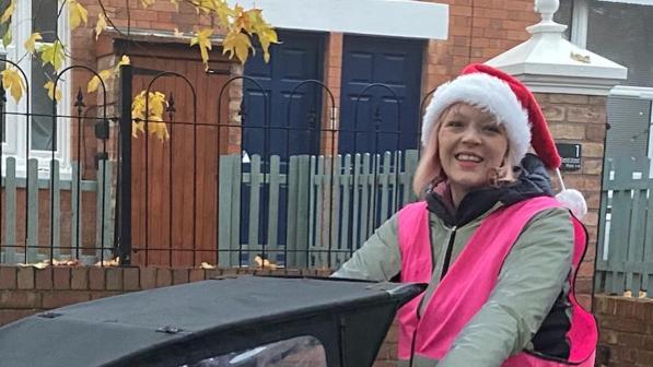 Katie Collier is riding a cargo bike in a pink high vis jacket and wearing a santa hat. At the front of the cargo bike there are children sitting under a black cover