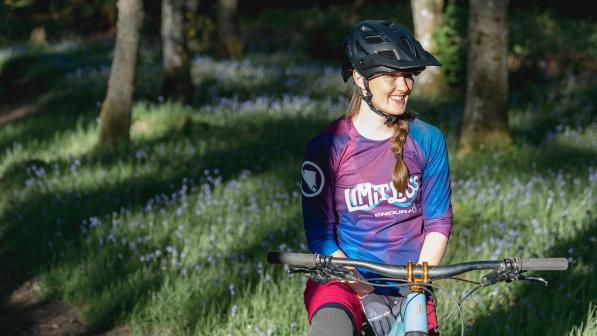 Katie May is sitting astride a mountain bike in a woodland. She is wearing a helmet and a Limitlass cycling jersey. Her blonde hair is in a plait. She is smiling.
