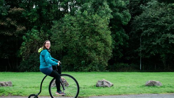 Katie Noble is smiling as she rides a penny farthing along a path. Behind her there is a grass lawn lined with trees
