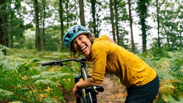 Katy Curd laughing while delivering mountain biking coaching in the Forest of Dean. She is leaning on the seat of a mountain bike. She is wearing a bright yellow top and cycling helmet.
