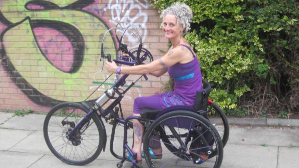 Kay Inckle is riding an adapted handcycle along a paved path. Behind her is a hedge and part of a brick wall with graffiti on it. She is wearing purple leggings and strappy top. She is smiling at the camera.