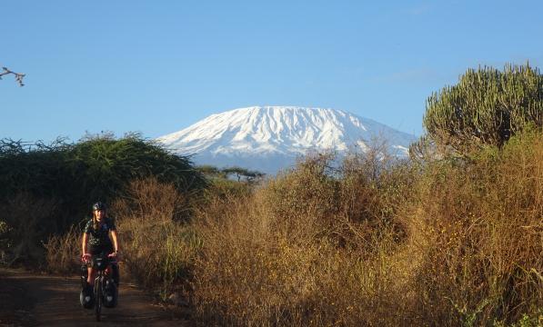 A woman on a loaded up touring bike with front and rear panniers is cycling on a dirt track through African countryside with Mount Kilimanjaro in the background