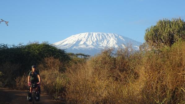 A woman on a loaded up touring bike with front and rear panniers is cycling on a dirt track through African countryside with Mount Kilimanjaro in the background