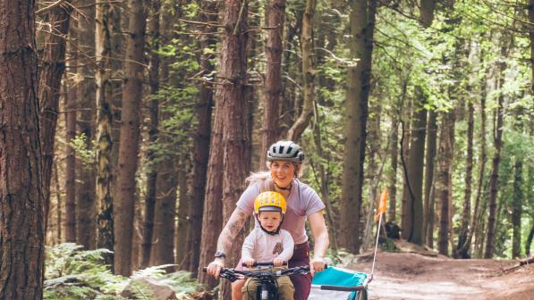 Kelly-Jayne Collinge is riding her bike on a woodland trail. Her with son Atlas is sat up front and she is pulling a trailer behind her. Mother and son are both wearing pink tops and helmets and both are smiling.