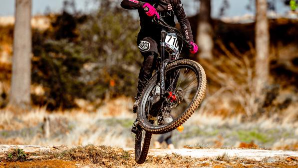 Kerry Wilson is performing a jump on a downhill off-road mountain bike track. She is wearing black mountain biking kit and a full face helmet. She has a race number (710) on her mountain bike handlebar.