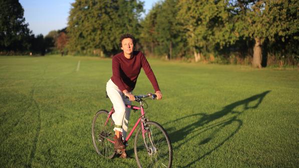Laura Di Giacomo on her pink bike in the park in Oxford. She is wearing a red long-sleeved T-shirt, white trousers and brown boots. She has short, curly brown hair.