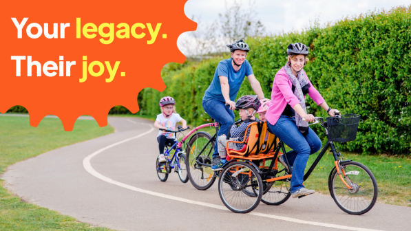 A family of five is cycling on a dedicated cycle path in a park. A woman is riding a trike with two youngsters on the back; a man is riding a bike with a tagalong attached and an older child riding. They are all wearing normal clothes with cycle helmets