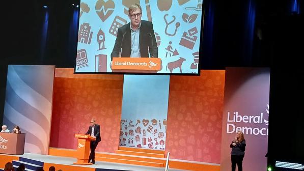 A man is standing in the centre of a stage decorated with orange panels, one of which has the Liberal Democrats logo. He's at a lectern and is giving speech. He's also shown on a big screen above him. A woman is standing to the right of the stage and there are two people sitting behind a desk to the left.