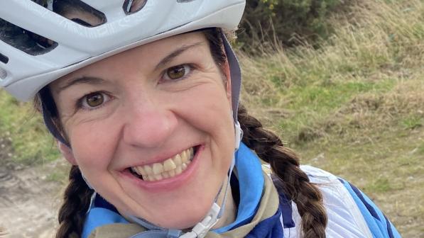 Lorna Breetzke shows off her blue and white Elgin Cycling Club kit. She has taken a selfie and is smiling at the camera. She has long brown hair in plaits and is wearing a cycle helmet. She’s out in the countryside and there’s a big gorse bush behind her.
