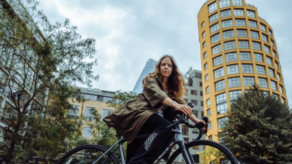 Lucy Poulden is sitting on a black road bike. She is on a road in front of a yellow tall building and is wearing a long green jacket and black trousers