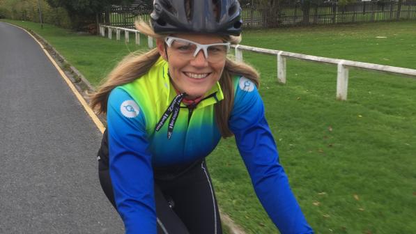 Lynn Bye rides her road bike togged up in Fat Lad at the Back cycling kit, a blue and yellow long-sleeved jersey and black leggings. She is wearing a helmet and cycling glasses; she has long blonde hair.