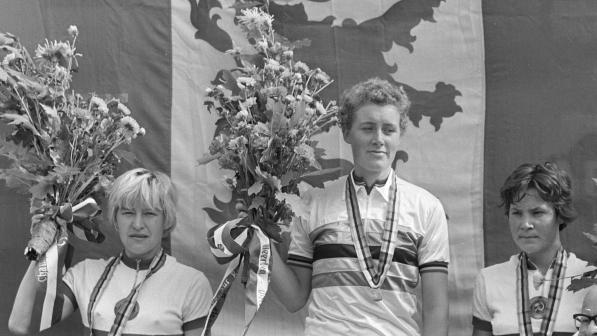 Three women are standing on a podium in front of a flag. They're holding flowers and have medals around their necks. They're wearing cycling sport kit