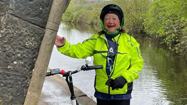 A woman is standing under a bridge over a canal. Her bike is leaning against the bridge. She is smiling and is wearing hi-vis cycling kit and a helmet