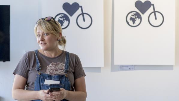 Marjan van de Weg at work at Bikes for Refugees Scotland. She is wearing denim dungarees and a taupe T-shirt. She has long blonde hair in a ponytail and sunglasses on her head. She is holding a smartphone. Behind her on the wall are two illustrations showing a cycle with a heart as the frame and a globe as the rear wheel.