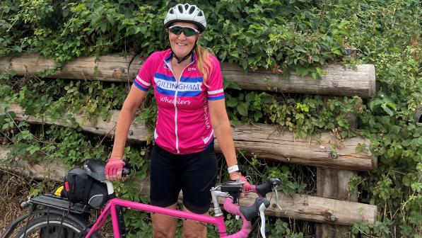 Mary Emerson-Reed is standing with a bright pink touring bike. She is wearing black shorts and a matching bright pink Gillingham Wheelers short-sleeved jersey. She has a helmet and cycling sunglasses. She is smiling at the camera.