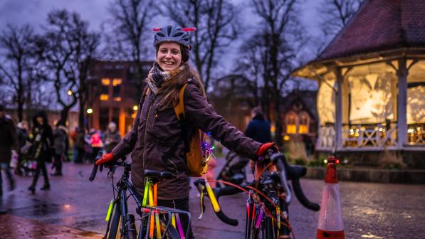 A woman smiles at the camera, holding a bike decorated with glow sticks on a street at twilight