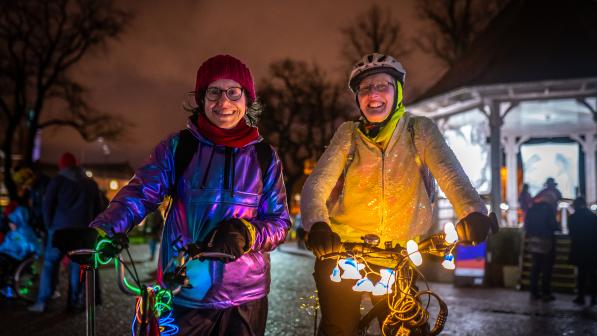 Two women stand smiling at the camera on a dark street with bikes lit up with fairy lights