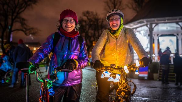 Two women stand smiling at the camera on a dark street with bikes lit up with fairy lights