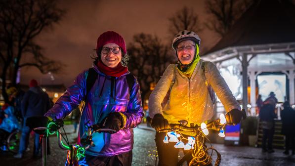 Two women stand smiling at the camera on a dark street with bikes lit up with fairy lights