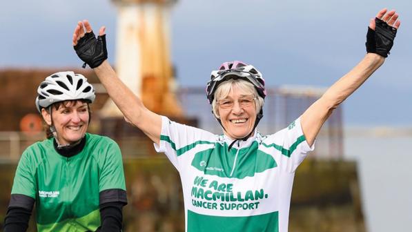 Mavis Paterson pictured at the finish line after completing her record-breaking LEJOG ride. She is smiling broadly and has both arms raised in the air. She is wearing a green t-shirt that reads ‘we are Macmillan Cancer Support’