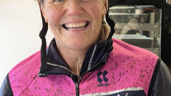 A smiling cyclist wearing a helmet and a mud-splattered Newbury Velo jersey exudes energy and enthusiasm. The background includes a display cabinet, suggesting a post-ride moment of reflection or celebration. Her expression captures the joy and passion she brings to cycling and her community.