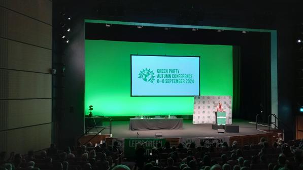 A man is standing on a stage to the right. A big green screen is displaying the Green Party logo and the words 'Green Party autumn conference 6-8 September 2024'