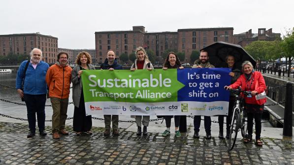 A group of nine people is standing in the rain on a cobbled path in front of a canal. They are holding a banner reading Sustainable Transport Alliance, it's time to get a shift on. One has a bike, one has an umbrella