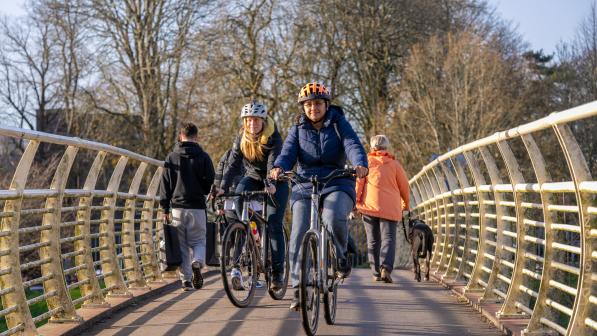 Two women cycle over a bridge towards the camera in autumn sunshine