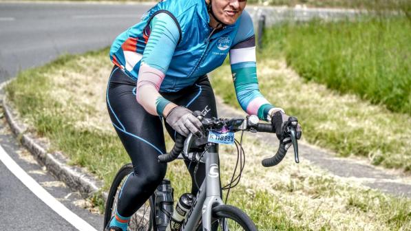 Nahida Hussain is riding a silver Cannondale road bike. She is wearing a Cycle Sisters cycling jersey and cycling leggings, as well as a helmet and cycling sunglasses. She has an event number fixed to her handlebar and is smiling.