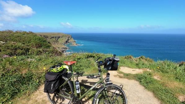 A loaded green Genesis bike is propped up on a gravel path on a gorse-covered clifftop with the sea in the background